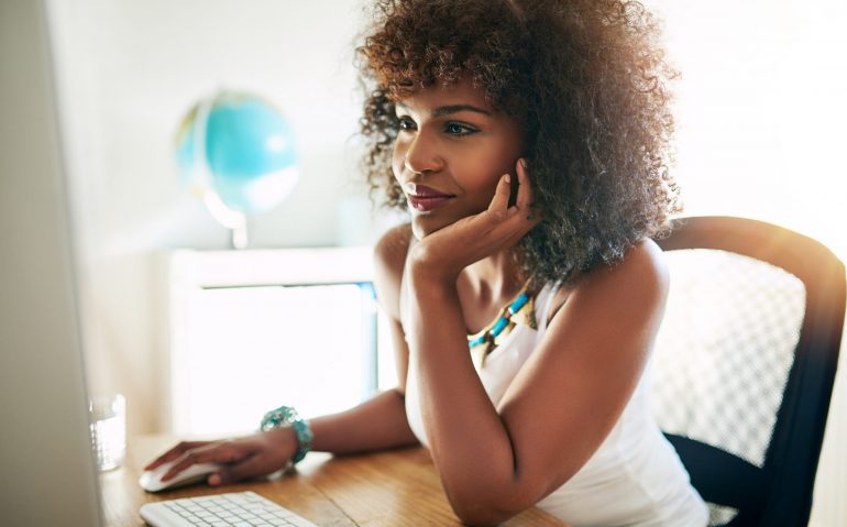 Young pretty afro-american woman using small business computer and smiling on blurred inside background.